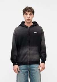 Νεαρός άνδρας που φοράει μαύρο φθαρμένο φούτερ με φερμουάρ και μπροστινές τσέπες, μαζί με φθαρμένο μπλε τζιν, που στέκεται μπροστά σε ένα απλό λευκό φόντο.