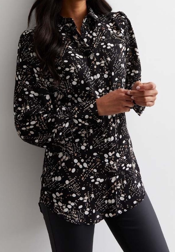 Femme portant un chemisier noir à manches longues boutonné, avec des motifs abstraits à pois blancs et un pantalon noir, debout devant un fond clair.