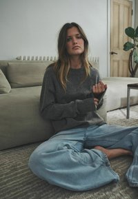 Sivý pletený sveter s dlhými rukávmi, doplnený o voľné modré džínsy. Prostredie tvorí neutrálna pohovka a drevené dvere.