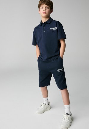Tumesinine polo-särk ja sobivad lühikesed püksid, valgest puuvillast; kaubamärgi logo on valge. Valged sneakerid heledate talladega ja valged sokid.