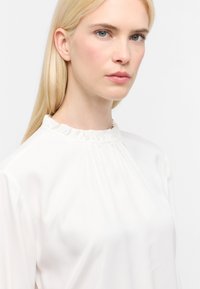 Blusa bianca con colletto alto arricciato, tessuto morbido, maniche lunghe e vestibilità ampia; presenta morbide pieghe per una silhouette elegante.
