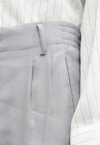 Pantalon gris clair taille haute avec passants de ceinture et plis, associé à une chemise blanche avec de fines rayures verticales noires.