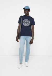 Marineblå t-shirt med cirkulært grafisk tryk, lyseblå jeans, hvide sneakers og en denim kasket. Modellen står med en crossbody taske.
