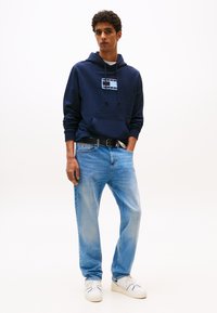 Noor mees, kes kannab mereväe sinist kapuutsiga pusa logoga, hele sinised teksapüksid ja valged tossud, seisab ühe käega taskus tavalise tausta ees.