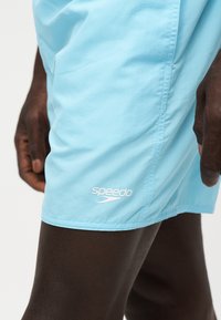 Gros plan d'une personne portant un short de bain Speedo bleu clair, montrant une partie de la jambe et de la main sur un fond uni.