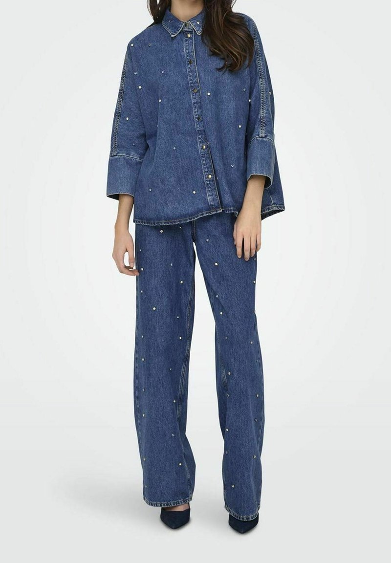 Femme portant une chemise en denim ample assortie et un jean à jambes larges décorés de petites perles blanches, associés à des chaussures noires pointues.