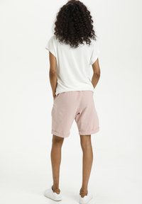Pantaloni corti rosa chiaro con risvolti, realizzati in un tessuto morbido, abbinati a una maglietta bianca a maniche corte, visti da dietro. Si indossano sneakers bianche.