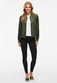 Giacca bomber di raso verde oliva con chiusura a zip, polsini e colletto a coste, indossata sopra una maglietta bianca e leggings neri. Scarpe nere open-toe.
