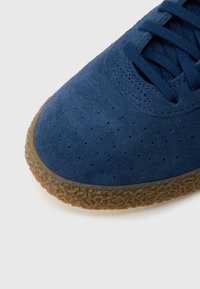 Σκούρος μπλε σουέτ αθλητικός παπούτσι με διάτρητες λεπτομέρειες, υφή από καουτσούκ σε σόλα χρώματος καφέ και κορδόνια στο ίδιο σκούρο μπλε χρώμα. Λείο πάνω μέρος.