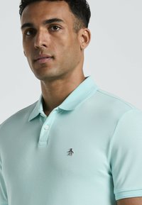 Lichtblauwe poloshirt van textuurstof, met een driebottonsluiting en een klein pinguïnlogo op de linkerborst.