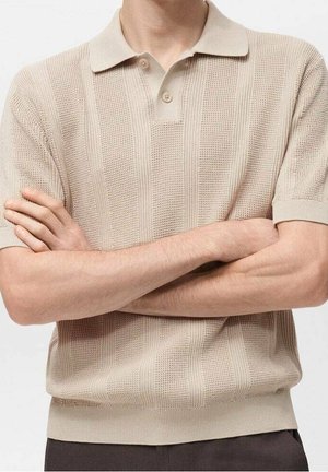 Mand iført beige kortærmet strikket polo med teksturerede lodrette striber, armene krydset, foran ensfarvet baggrund.