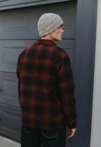 Junger Mann trägt eine graue Strickmütze, eine rot-schwarz karierte Jacke und dunkle Jeans und steht seitlich vor einer grauen Garagentür.