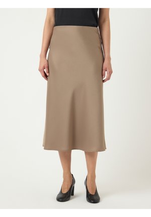 Kvinde iført en beige midinederdel, sort top og sorte højhælede sko står mod en hvid baggrund.