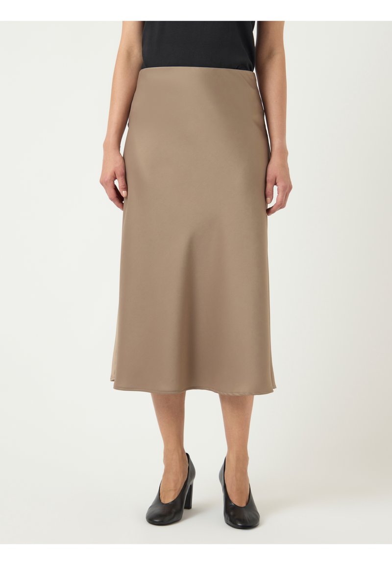 Femme portant une jupe midi beige, un top noir et des chaussures à talons hauts noires, debout devant un fond blanc.
