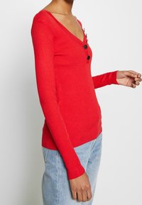 Haut rouge côtelé à manches longues avec un col en V profond et trois boutons noirs ; coupe ajustée avec tissu texturé et design minimaliste.