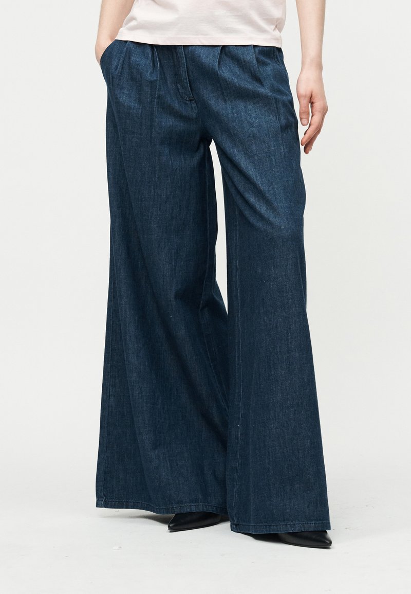 Femme portant un pantalon en jean bleu foncé à jambes larges et des chaussures noires à bouts pointus, avec une main dans la poche, debout devant un fond uni.