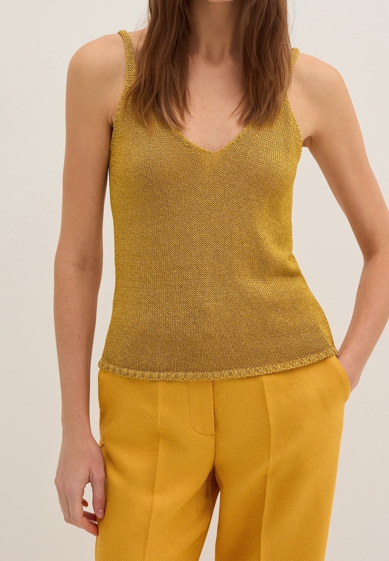 Femme portant un haut sans manches en maille jaune moutarde et un pantalon taille haute assorti, une main dans la poche, sur un fond uni.