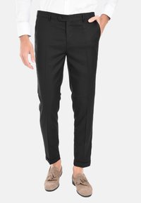 Pantaloni neri su misura con fronte piatto, taglio dritto, passanti per cintura e chiusura a bottone singolo. Abbinati a mocassini di colore chiaro.