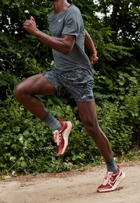 Biegacz w szarym t-shircie i szortach w kamuflaż biegnie po dirtowej ścieżce otoczonej zielonymi liśćmi.