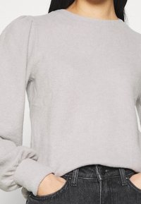 Svetle šedý pletený svetr s ribbed texturou, kulatým výstřihem a nafouknutými rukávy, v kombinaci s tmavými džínami s vysokým pasem.