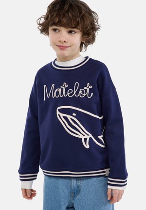 Enfant aux cheveux bruns et bouclés portant un pull marine avec le texte "Matelot" et un motif de baleine, associé à un jean bleu clair, debout devant un fond blanc.