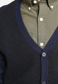 Mörk kofta med en texturerad stickning, marinblå kant och två svarta knappar, lager på lager över en olivgrön skjorta med krage.