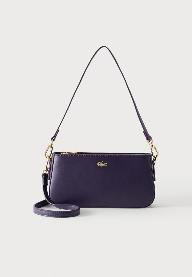 Sac à main rectangulaire violet foncé avec fermeture éclair dorée, logo crocodile doré et deux fines bandoulières amovibles sur fond blanc.