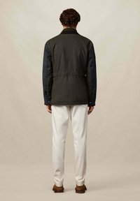 Muškarac s kovrčavom kosom stoji okrenut leđima, odjeven u tamnozeleni kaput, bijele hlače i smeđe cipele, na jednostavnoj bež pozadini.