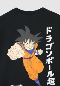 Czarna koszulka z grafiką postaci w pomarańczowym stroju, w dynamicznej pozie, z wyrazistym japońskim napisem z boku.