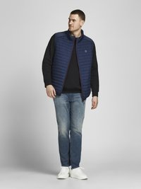 Navy quilted vest with high collar over black long-sleeve shirt, paired with light blue jeans and white sneakers. Simple design and modern fit.