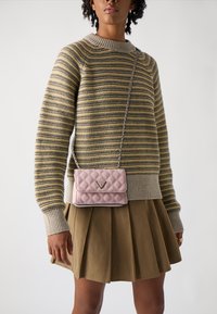Gestepptes rosa Handtasche mit einem dreieckigen Logo, getragen an einer silbernen Kette. Das Modell trägt einen gestreiften Pullover und einen plissierten Rock.