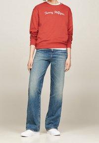 Roter Pullover mit der Aufschrift "Tommy Hilfiger" in Weiß bestickt, kombiniert mit lockeren hellblauen Jeans und weißen Sneakern. Glattes Material, lässiger Look.