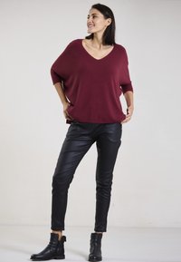 Bordeauxfarbener, locker sitzender Pullover mit V-Ausschnitt, kombiniert mit schwarzen Kunstlederhosen und schwarzen Knöchelstiefeln, vor einem hellen Hintergrund.