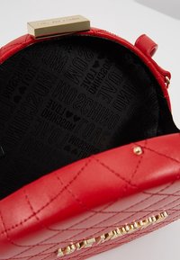 Červená prošívaná kabelka se hladkým povrchem, zlatým kováním a černým interiérem se vzorem opakujících se log. 