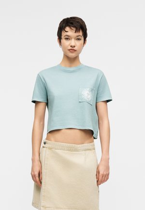 Fiatal nő rövid sötét hajjal, világoskék, hasat takaró pólót visel, melynek zsebében nap grafika látható, valamint bézs színű, átkötős szoknyát.