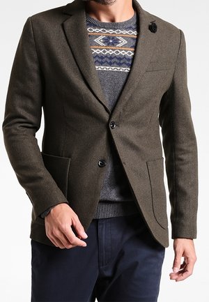 Hombre con un blazer verde oliva sobre un suéter gris con patrón y pantalones azul marino, con un pin de solapa negro en el blazer.