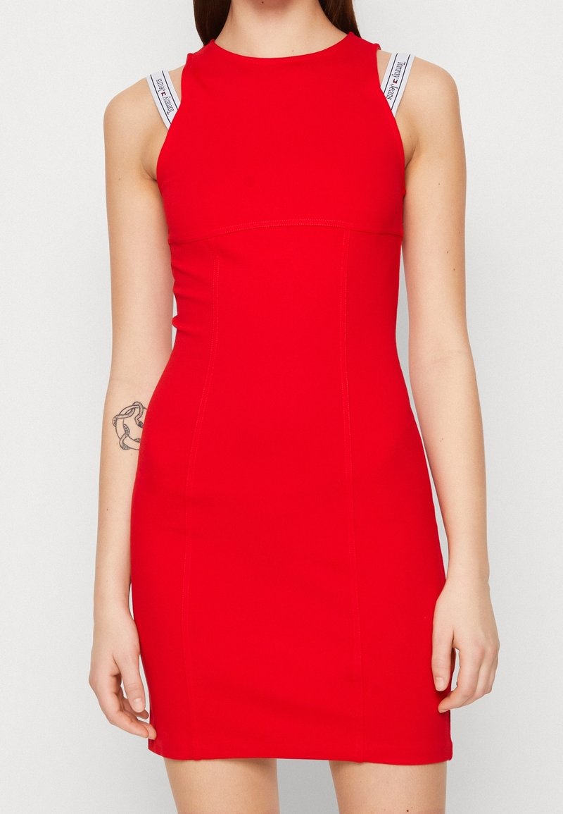 Femme portant une robe rouge ajustée sans manches avec des bretelles de soutien-gorge blanches visibles avec un texte noir.