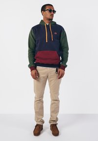 Hoodie met marineblauwe, groene en bordeauxrode kleurvakken, oranje trekkoorden en een kangoeroezak. Combinatie met beige broek en bruine schoenen.