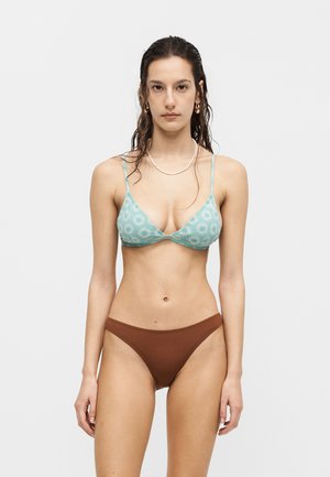 Kobieta z mokrymi ciemnymi włosami, ubrana w niebieski kwiatowy top bikini, brązowe majtki bikini, złote kolczyki koła oraz biały naszyjnik z koralików, stojąca.