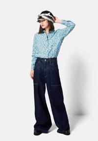 Bloemenprint blauw overhemd met knopen, donkerblauwe wijde hoge taille spijkerbroek, zwarte plateau schoenen en een decoratieve zwart-witte hoed.