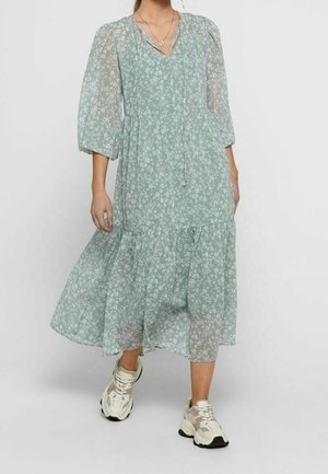 Light green floral dress with long sheer sleeves, v-neck, and tiered skirt; paired with white and beige athletic shoes.