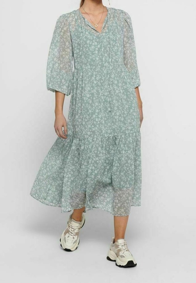Robe florale vert clair avec manches longues transparentes, col en V et jupe à volants ; associée à des chaussures de sport blanches et beiges.