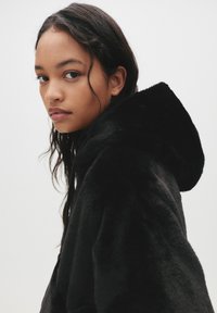 Črna jakna iz umetnega krzna z kapuco, ki ima mehak material in sproščen kroj, z obsežno kapuco in subtilnim šivom.
