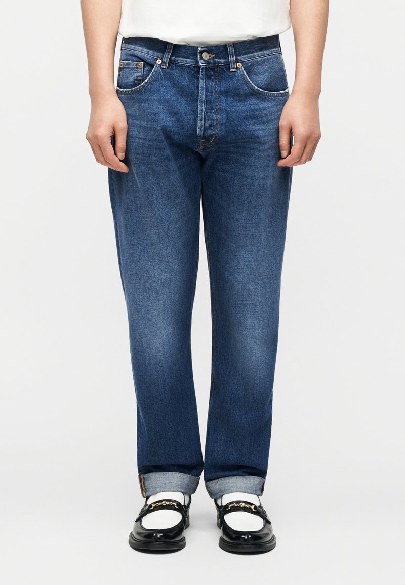 Άτομο που φοράει γαλάζια τζιν τζάκετς που έχουν  γυριστεί, λευκές κάλτσες και μαύρα μοκασίνια με χρυσή αλυσίδα, στέκεται μπροστά σε λευκό φόντο.