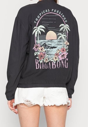 Kvinde iført sort jakke med farverig tropisk solnedgang, palmer, blomster og teksten "Tropical Feelings Billabong" på ryggen.