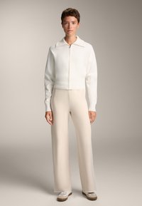 Helle, zip-festing Strickjacke mit Kragen, kombiniert mit beigen, weiten Hosen. Das Outfit zeichnet sich durch eine weiche Textur und ein minimalistisches Design aus.