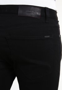 Vaqueros de denim negro con un parche de cuero con el logo HUGO en la cintura y el bolsillo trasero. Detalles de costura en los bolsillos. Corte estándar.