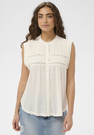 Žena s dlhými vlnitými vlasmi má na sebe bezrukávovú bielu blúzku so zapínaním na gombíky a textúrovanými zvislými pruhmi a svetlomodrú rifľovú sukňu s roztrhaným vzhľadom.