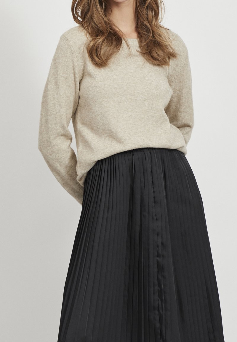 Beige gebreide trui met lange mouwen, gecombineerd met een zwarte geplooide rok met een gladde textuur en verticale plooien.