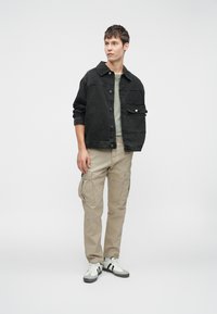 Chaqueta de denim negra con cierre de botones, pantalones de cargo beige con bolsillos laterales y zapatillas blancas con detalles negros. Fondo neutro.